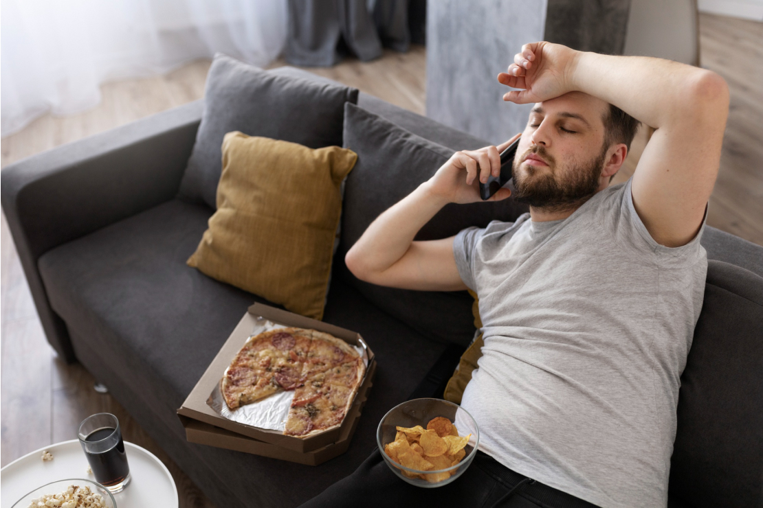 A tired man on a couch with pizza and chips, symbolizing hunger from poor sleep.