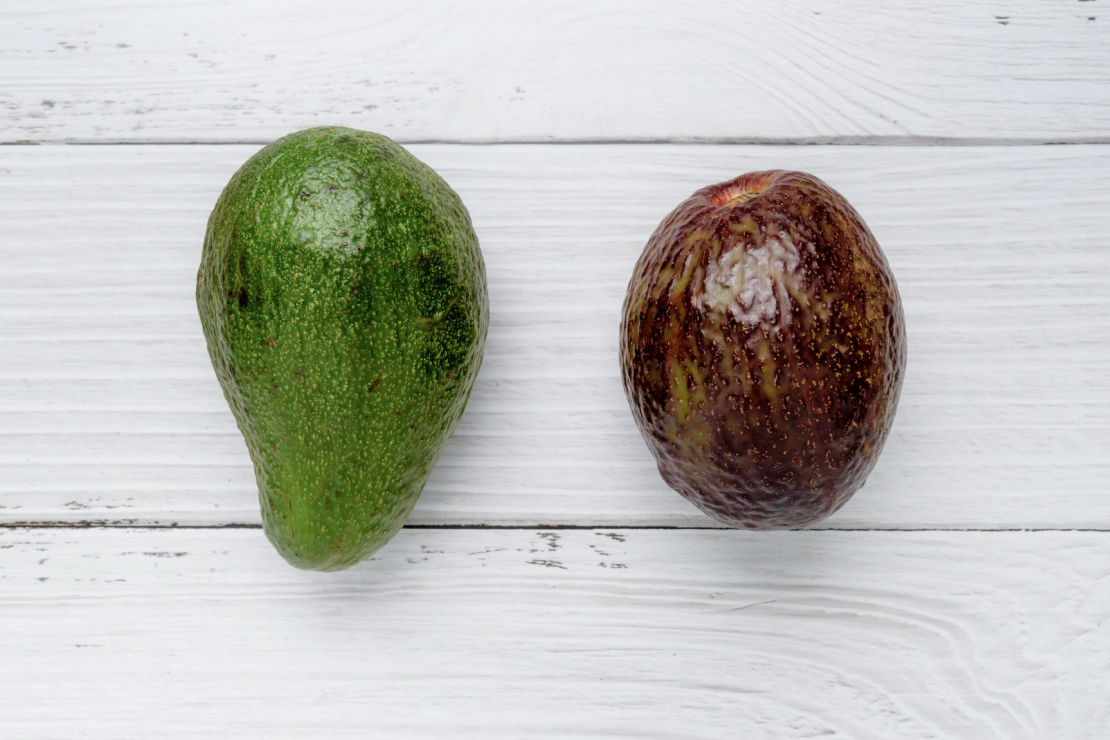 A side-by-side comparison of a green organic avocado and a darker non-organic avocado on a white wooden surface.