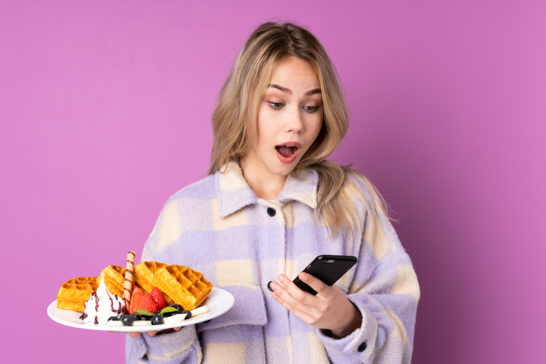 A person looking surprised at their phone while holding a plate of waffles, symbolizing the discovery of hidden sugars with AI.