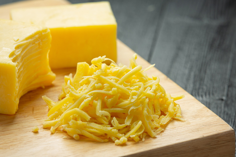 Block and shredded cheddar cheese on a wooden board, ready for cooking.